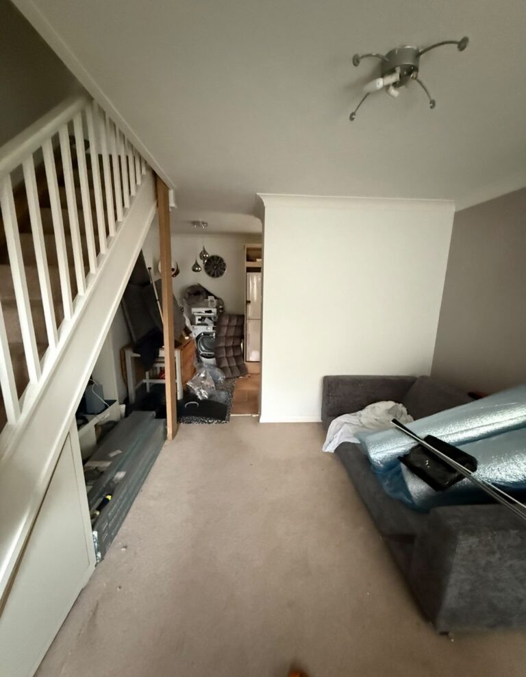 Max's living room before: worn beige carpet, bare walls and boxes of flooring waiting under the stairs